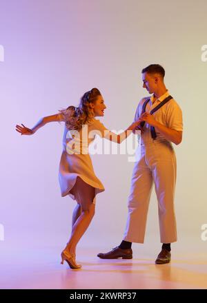 Danse incendiaire. Couple émotionnel de danseurs en tenues rétro dansant social danses isolées sur fond de couleur lilas dégradé dans la lumière de néon Banque D'Images