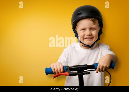 jeune garçon souriant avec un casque debout sur un scooter de coup sur fond jaune avec espace de copie Banque D'Images