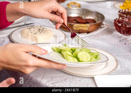 Femme tenant et servant une assiette de concombre tranché. Vue rapprochée. Saucisse appelée sucuk en arrière-plan. Fromage blanc et confiture sur la table. Banque D'Images