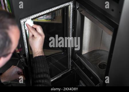 Homme nettoyant la cendre sur le verre d'une pastille ou d'un poêle à bois avec une serviette en papier et de l'eau chaude Banque D'Images