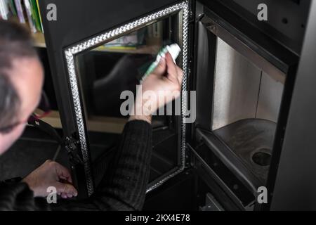 Homme nettoyant la cendre sur le verre d'une pastille ou d'un poêle à bois avec une serviette en papier et de l'eau chaude Banque D'Images