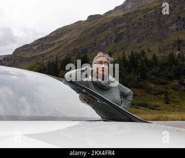 Une jeune femme aux cheveux gris dans un chandail chaud se tient près d'une voiture dans une vallée alpine Banque D'Images