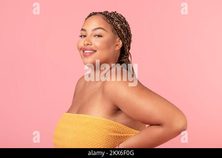 Portrait d'une femme afro-américaine obèse enveloppée d'une serviette souriante à l'appareil photo, posant sur fond rose studio Banque D'Images