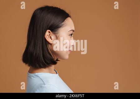 Jeune femme asiatique souriant en posant dans le profil isolé sur fond beige Banque D'Images