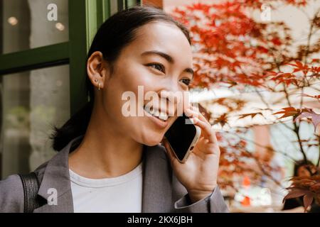 Jeune femme d'affaires asiatique souriant et parlant au téléphone portable tout en se tenant dans un bâtiment dans la rue Banque D'Images