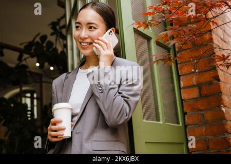 Jeune femme d'affaires asiatique souriant et parlant au téléphone portable tout en se tenant près de la porte dans la rue Banque D'Images