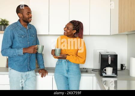 Joyeux jeune couple noir qui boit du café et bavarde à l'intérieur de la cuisine, les conjoints appréciant une boisson chaude à la maison Banque D'Images