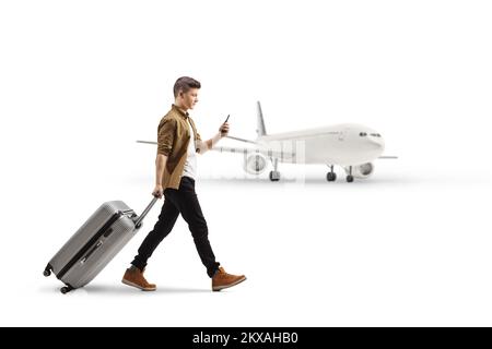 Photo pleine longueur d'un jeune homme avec un smartphone tirant une valise devant un avion isolé sur fond blanc Banque D'Images