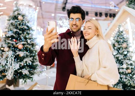 Portrait d'un jeune couple amical en train de parler en regardant un téléphone intelligent passer un appel vidéo, en donnant des salutations en ligne du nouvel an debout dans le hall de la fête Banque D'Images