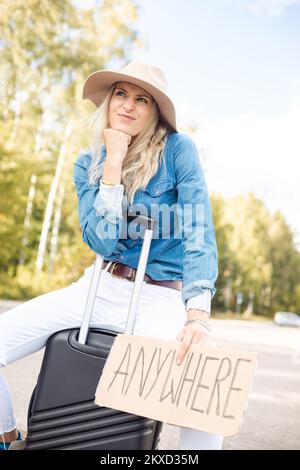 Une femme qui fait la gestuelle arrête de passer la voiture assise sur une valise avec une affiche en carton dans la forêt sur le bord de la route, de la vue d'en dessous. Dame en chapeau échapper de la ville à partir Banque D'Images