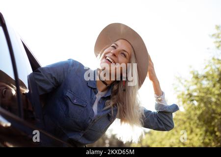 Une femme rêveuse quitte joyeusement la ville en se tenant la tête hors de la fenêtre ouverte de la voiture et en regardant vers le ciel. Jeune femme blonde dans le chapeau avec l'humeur heureuse aller à l'auto Banque D'Images