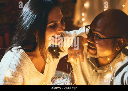 Heureux couple interracial célébrant le nouvel an à la maison ensemble, assis guirlande à proximité l'un de l'autre Banque D'Images