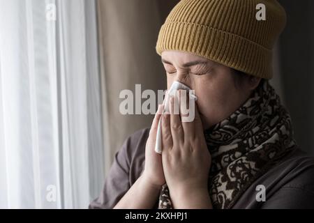 Malade brune femme dans le chapeau et le foulard éternue couvrant le nez avec la sensation de serviette. Banque D'Images