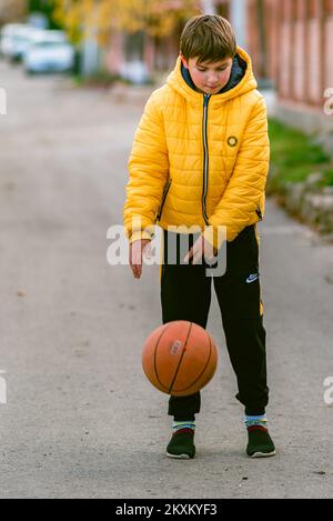 Jeune garçon tenant entre les mains Sport de basket-ball Jeune petit sportif caucasien enfant jouant à la tête de la rue de balle orange dribbling jaune veste matelassée o Banque D'Images