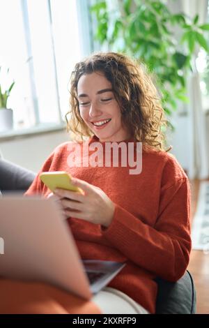 Bonne jeune femme se reposant sur un canapé en utilisant le téléphone dans un salon confortable, vertical. Banque D'Images