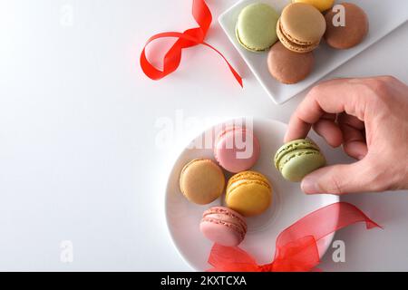 Dégustation à la main de macarons français d'une assiette blanche aux saveurs différentes sur une table blanche aux noeuds rouges. Vue de dessus. Banque D'Images