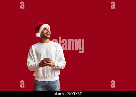 Joyeux homme arabe portant un chapeau de père Noël tenant un smartphone et regardant de côté Banque D'Images