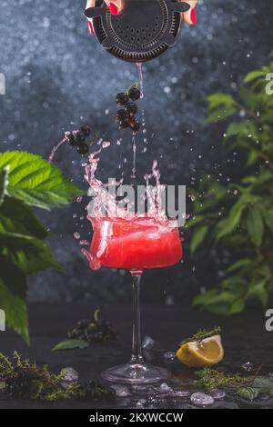 Élégant verre de Black Currant Gin Sour cocktail ou Mockttails avec des éclaboussures entourées d'ingrédients sur une surface de table sombre. Boisson rafraîchissante prête pour Banque D'Images