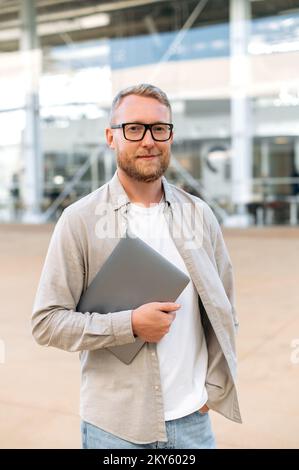 Photo verticale d'un homme caucasien à barbe positive et confiante, avec des lunettes, dans un vêtement décontracté, un programmeur, seo, tient un ordinateur portable dans sa main, regarde la caméra, sourit amical Banque D'Images