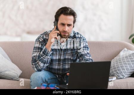 Triste pensif millennium européen gars dans les vêtements de maison avec les appels de chaume par téléphone, regardez l'ordinateur Banque D'Images