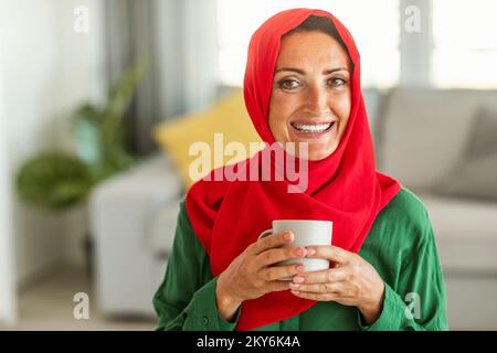 Portrait d'une heureuse femme islamique d'âge moyen dans le hijab assis dans le salon avec une tasse de tisane et souriant à l'appareil photo Banque D'Images
