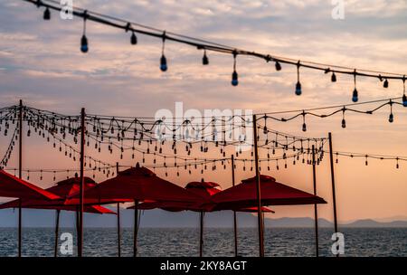 Belle scène de coucher de soleil avec de nombreuses plages de parasols rouges et des ampoules de fête accrochent sur l'air, ciel coloré de coucher de soleil. Banque D'Images