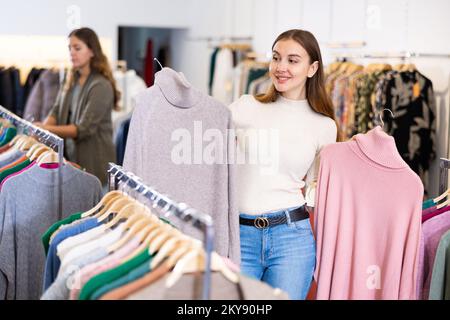 Bonne jeune femme qui choisit entre les pull-overs roses et beiges dans un magasin de vêtements Banque D'Images