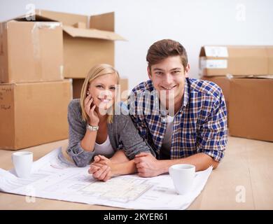 Décider de la disposition de leur nouvel appartement. Un jeune couple allongé sur le sol avec les plans de leur nouvel appartement et des boîtes derrière eux. Banque D'Images