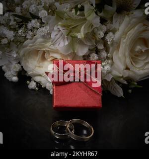 Deux anneaux de différentes tailles, une boîte cadeau rouge se trouve sur le fond d'un bouquet de fleurs. La photographie dans une obscurité. Gros plan. Mise au point sélective, flou Banque D'Images