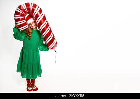 Fille de Noël tenant un gros ballon de canne dans les mains, portant dans la robe verte, chapeau de père noël et chaussettes chaudes rouges pour la fête du nouvel an Banque D'Images