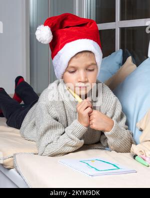 Une liste de souhaits pour un miracle de Noël. Un enfant sérieux concentré dans un chapeau de Santa écrit une liste de souhaits ou une lettre à Santa à la maison. Le garçon tire un téléphone. A ch Banque D'Images