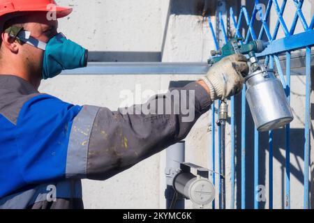 Un peintre industriel en respirateur et casque peint la clôture métallique à partir d'un pistolet pulvérisateur. Peinture de la clôture le jour d'été. Flux de travail authentique. Travailleur professionnel en combinaison... Banque D'Images