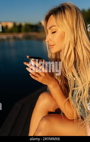 Portrait vertical de la jeune femme blonde calme avec les yeux fermés boire du café chaud de la coupe de thermos assis sur la plage l'été matin ensoleillé. Banque D'Images