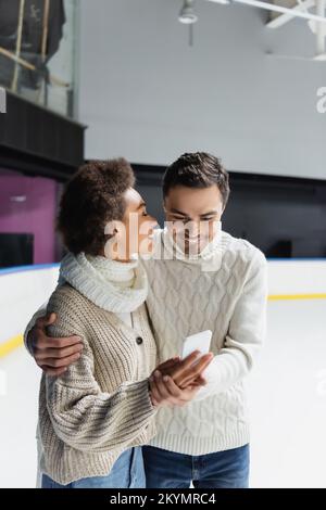 Couple interracial souriant qui se serre et utilise un smartphone sur une patinoire Banque D'Images