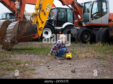 garçon jouant avec un bulldozer jouet devant plusieurs grosses pelles hydrauliques sur roues avec d'énormes godets en métal. Le passe-temps du garçon pour les équipements de construction Banque D'Images