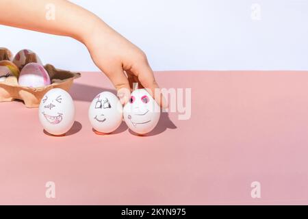 L'enfant peint des oeufs blancs et les met sur un fond rose et blanc. Visages amusants et romantiques dessinés avec un marqueur. Concept de vacances de Pâques. Banque D'Images