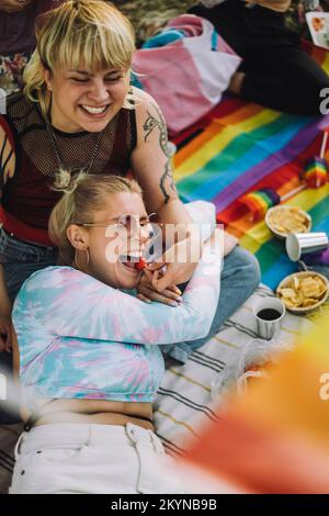 Bonne personne non binaire nourrissant la fraise à une amie allongée sur une couverture de pique-nique Banque D'Images
