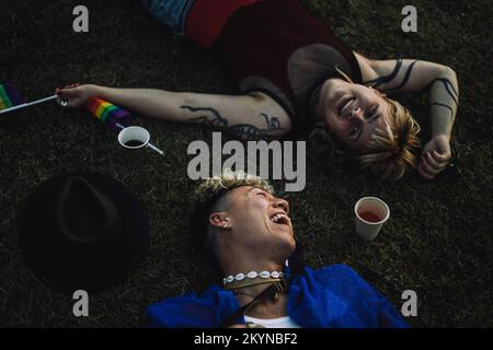 Jeune homme gai couché par un ami non binaire sur l'herbe au parc pendant le coucher du soleil Banque D'Images