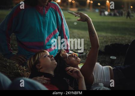 Jeune femme prenant selfie avec une personne non binaire tout en étant couché sur les genoux d'un ami pendant le coucher du soleil Banque D'Images