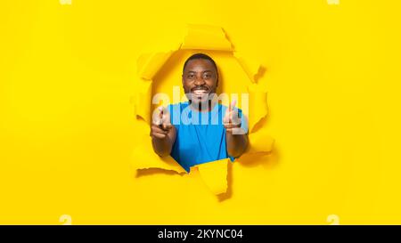 Et qu'en est-il de vous ? Heureux afro-américain homme pointant les doigts vers la caméra, brisant à travers le trou dans le papier jaune Banque D'Images