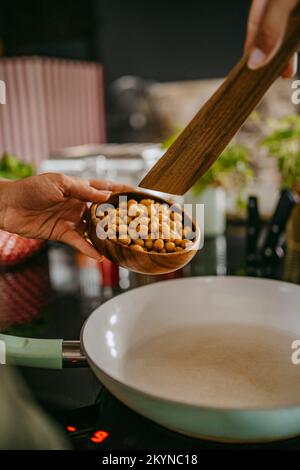 Les mains du chef féminin ajoutant le pois chiche dans une casserole à l'aide d'une cuillère en bois dans la cuisine du studio Banque D'Images