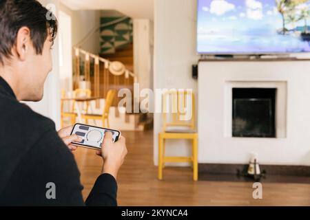 Homme utilisant l'application Smart Home sur un téléphone mobile tout en regardant la télévision dans le salon à la maison Banque D'Images