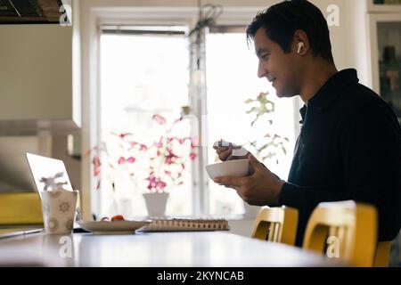 Vue latérale d'un travailleur indépendant qui prend le petit-déjeuner tout en participant à une vidéoconférence sur ordinateur portable à la maison Banque D'Images