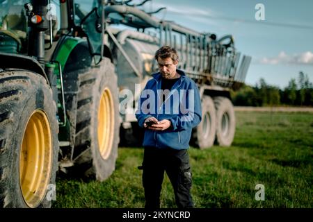 Agriculteur mature utilisant un smartphone debout contre le tracteur dans les champs Banque D'Images