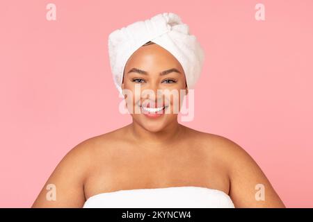 Portrait de beauté du corps positif femme noire debout enveloppée dans une serviette sur fond rose et souriant à l'appareil photo Banque D'Images