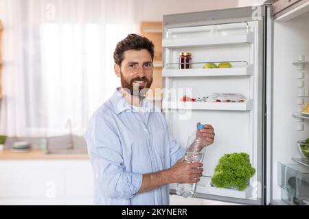 Gai mature caucasien mâle avec la barbe dans la chemise ouvre la porte du réfrigérateur, prend une bouteille d'eau à l'intérieur de la cuisine Banque D'Images