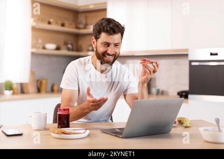 Souriant d'âge moyen homme caucasien avec la barbe en t-shirt blanc mange sandwich, a appel vidéo, se gesticulent Banque D'Images