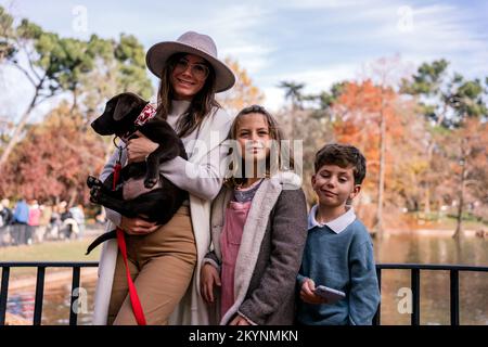 Bonne maman en vêtements d'extérieur embrassant le Labrador Retriever chiot avec laisse et sourire en se tenant près des enfants sur le pont au-dessus de l'étang le week-end Banque D'Images