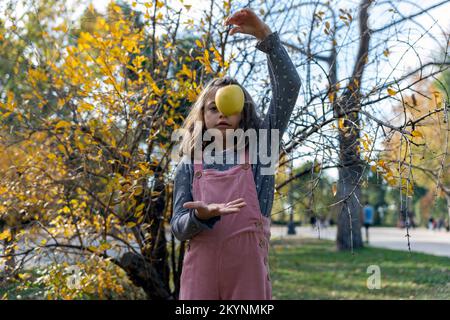Jolie fille dans des vêtements décontractés qui laisse tomber la pomme fraîche à la main tout en se tenant près d'un arbuste avec des feuilles jaunes le jour de l'automne dans le parc Banque D'Images
