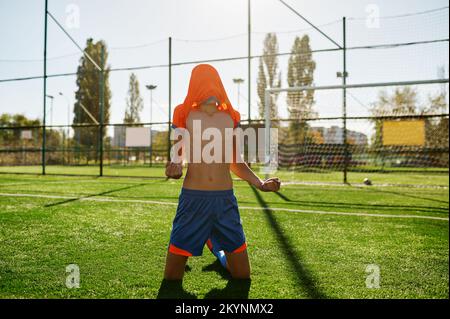 Joueur de football de garçon émotionnel avec t-shirt sur la tête se réjouissant but ou gagner Banque D'Images